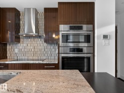 Kitchen featuring double oven, modern cabinets, wall chimney range hood, backsplash, and light stone countertops - 