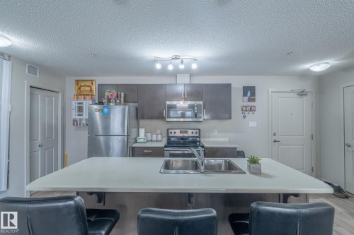 Kitchen with stainless steel appliances, a breakfast bar area, light countertops, a center island with sink, and a textured ceiling - 107 142 Ebbers Boulevard, Edmonton, AB - Indoor Photo Showing Kitchen With Double Sink With Upgraded Kitchen