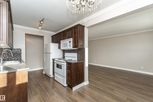 Kitchen with white appliances, tasteful backsplash, dark brown cabinets, ornamental molding, and a chandelier - 102 10735 81 Avenue, Edmonton, AB - Indoor Photo Showing Kitchen With Double Sink
