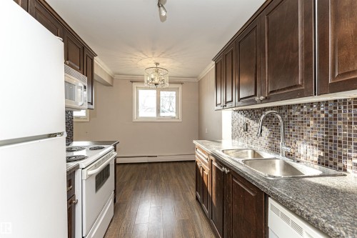 Kitchen featuring white appliances, dark brown cabinetry, ornamental molding, decorative backsplash, and a chandelier - 102 10735 81 Avenue, Edmonton, AB - Indoor Photo Showing Kitchen With Double Sink