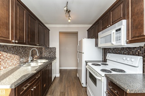 Kitchen with white appliances, dark brown cabinetry, crown molding, dark countertops, and dark wood-style floors - 102 10735 81 Avenue, Edmonton, AB - Indoor Photo Showing Kitchen With Double Sink