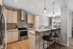 Kitchen with light brown cabinets, stainless steel appliances, hanging light fixtures, backsplash, and wall chimney range hood - 