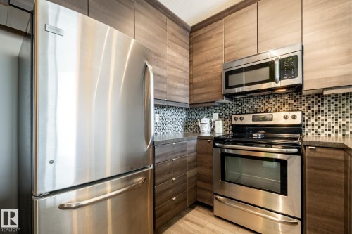 Kitchen featuring stainless steel appliances, dark countertops, decorative backsplash, light wood-style floors, and modern cabinets - 407 2590 Anderson Way, Edmonton, AB - Indoor Photo Showing Kitchen