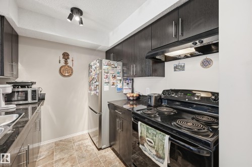 Kitchen featuring stainless steel appliances, under cabinet range hood, a textured ceiling, and dark cabinetry - 207 1080 Mcconachie Boulevard, Edmonton, AB - Indoor Photo Showing Kitchen