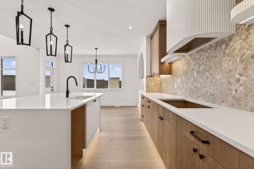 Two tone kitchen featuring light stone countertops, backsplash, light wood-style flooring, a kitchen island with sink, and black electric cooktop - 9 Elwyck Gate, Spruce Grove, AB - Indoor Photo Showing Kitchen With Upgraded Kitchen