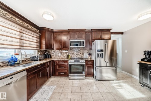 Kitchen featuring stainless steel appliances, dark wood finish cabinetry, light stone countertops, and light tile patterned floors - 12203 147 Avenue, Edmonton, AB - Indoor Photo Showing Kitchen With Stainless Steel Kitchen With Double Sink With Upgraded Kitchen