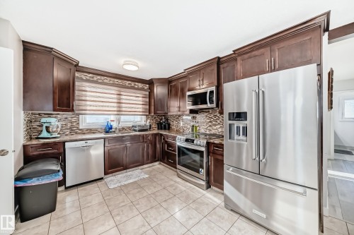 Kitchen featuring stainless steel appliances, dark wood finish cabinets, light tile patterned flooring, light stone countertops, and decorative backsplash - 12203 147 Avenue, Edmonton, AB - Indoor Photo Showing Kitchen With Stainless Steel Kitchen With Upgraded Kitchen