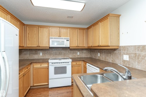 Kitchen featuring white appliances, decorative backsplash, a textured ceiling, wood finished floors, and light wood finish cabinetry - 112 260 Sturgeon Road, St. Albert, AB - Indoor Photo Showing Kitchen With Double Sink
