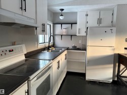 Kitchen with white appliances, dark countertops, white cabinets, under cabinet range hood, and hanging light fixtures - 