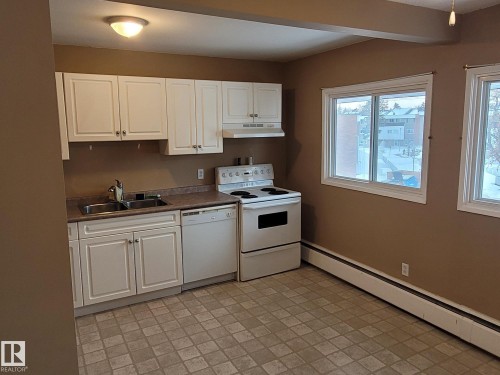 Kitchen featuring a baseboard radiator, white appliances, dark countertops, and white cabinets - 304A 2908 116A Avenue, Edmonton, AB - Indoor Photo Showing Kitchen With Double Sink