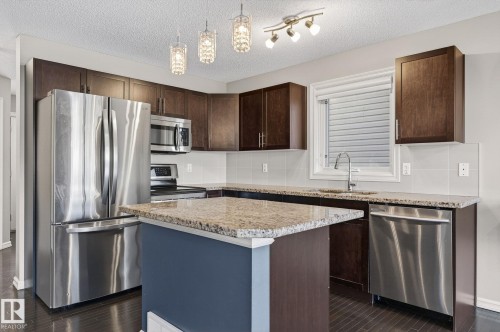 Kitchen with stainless steel appliances, a center island, dark wood finish cabinetry, hanging light fixtures, and light stone countertops - 2338 Casselman Crescent, Edmonton, AB - Indoor Photo Showing Kitchen With Stainless Steel Kitchen With Upgraded Kitchen
