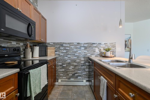 Kitchen featuring black appliances, wood finish cabinets, decorative light fixtures, a baseboard heating unit, and tasteful backsplash - 411 1619 James Mowatt Trail, Edmonton, AB - Indoor Photo Showing Kitchen With Double Sink