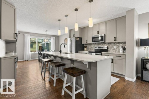 Kitchen with gray cabinetry, decorative light fixtures, a kitchen bar, stainless steel appliances, and a center island with sink - 3351 Orchards Link, Edmonton, AB - Indoor Photo Showing Kitchen With Upgraded Kitchen
