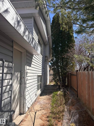 Exterior siding with multiple windows and a side entrance, bordered by a wooden fence and established trees - 5128 52 Street, Barrhead, AB - Outdoor