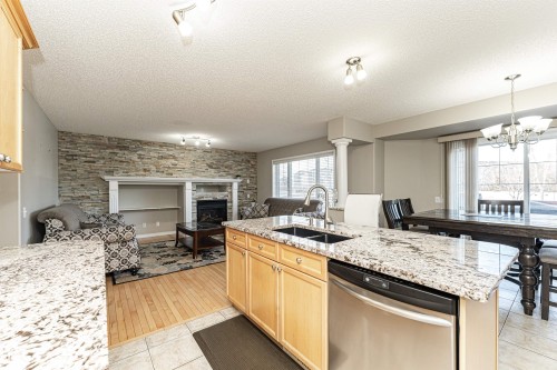 Kitchen featuring dishwasher, light wood finish cabinets, light stone countertops, a stone fireplace, and a textured ceiling - 192 Keyport Circle, Leduc, AB - Indoor Photo Showing Kitchen With Fireplace With Double Sink