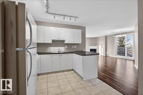 Kitchen with white appliances, white cabinets, a fireplace, a peninsula, and light tile patterned floors - 402 10649 Saskatchewan Drive, Edmonton, AB - Indoor Photo Showing Kitchen With Double Sink