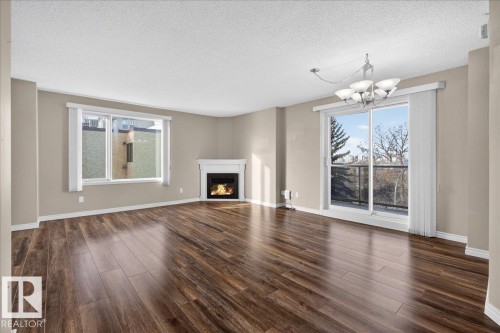 Unfurnished living room with a lit fireplace, dark wood-style flooring, a textured ceiling, and hanging lights - 402 10649 Saskatchewan Drive, Edmonton, AB - Indoor Photo Showing Living Room With Fireplace