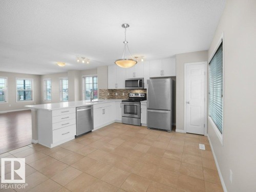 Kitchen featuring stainless steel appliances, white cabinetry, open floor plan, light tile patterned flooring, and a textured ceiling - 15 7385 Edgemont Way, Edmonton, AB - Indoor Photo Showing Kitchen
