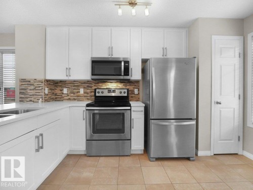 Kitchen with stainless steel appliances, white cabinetry, light tile patterned floors, a textured ceiling, and light stone counters - 15 7385 Edgemont Way, Edmonton, AB - Indoor Photo Showing Kitchen With Double Sink