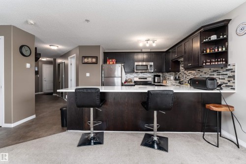 Kitchen with stainless steel appliances, a breakfast bar, dark wood finish cabinetry, a peninsula, and open shelves - 325 1520 Hammond Gate, Edmonton, AB - Indoor Photo Showing Kitchen With Upgraded Kitchen