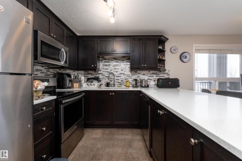 Kitchen with stainless steel appliances, open shelves, dark wood finish cabinetry, light stone counters, and rail lighting - 325 1520 Hammond Gate, Edmonton, AB - Indoor Photo Showing Kitchen With Double Sink