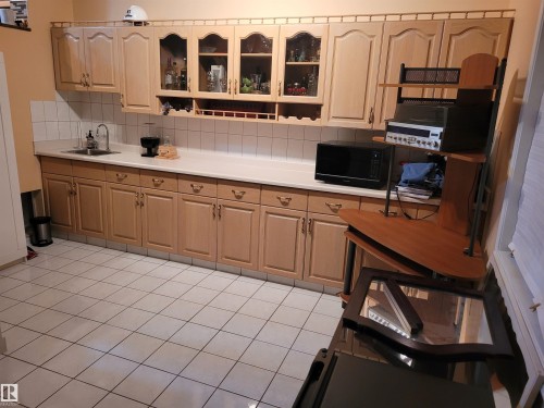 Kitchen featuring light countertops, decorative backsplash, glass insert cabinets, light tile patterned floors, and light wood finish cabinetry - 2812 35 Street Nw, Edmonton, AB - Indoor Photo Showing Kitchen With Double Sink