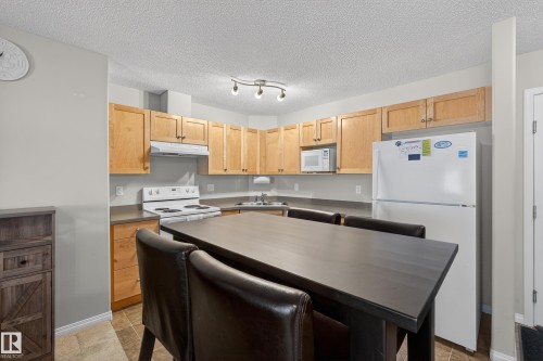 Kitchen featuring light wood finish cabinetry, white appliances, dark countertops, and a textured ceiling - 131 1520 Hammong Gate, Edmonton, AB - Indoor Photo Showing Kitchen With Double Sink