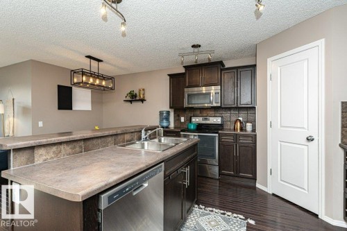 Kitchen with a kitchen island with sink, stainless steel appliances, dark wood finish cabinetry, tasteful backsplash, and dark wood-style floors - 6649 Cardinal Road Sw, Edmonton, AB - Indoor Photo Showing Kitchen With Double Sink