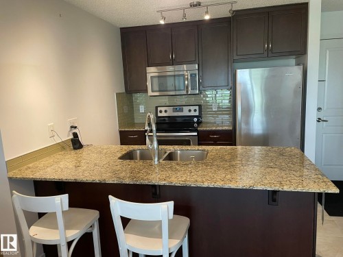 Kitchen featuring a breakfast bar, dark wood finish cabinets, stainless steel appliances, light stone counters, and a textured ceiling - 207 7021 South Terwillegar Drive, Edmonton, AB - Indoor Photo Showing Kitchen With Stainless Steel Kitchen With Double Sink With Upgraded Kitchen