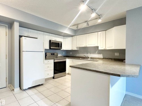 Kitchen with appliances with stainless steel finishes, a peninsula, dark countertops, white cabinetry, and light tile patterned flooring - 1402 10909 103 Avenue, Edmonton, AB - Indoor Photo Showing Kitchen With Double Sink