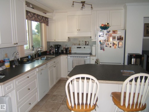 Kitchen with white appliances, decorative backsplash, white cabinetry, light tile patterned flooring, and ornamental molding - 4807 54 Avenue, Two Hills, AB - Indoor Photo Showing Kitchen With Double Sink