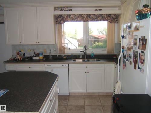 Kitchen featuring white appliances, dark countertops, white cabinets, decorative backsplash, and light tile patterned floors - 4807 54 Avenue, Two Hills, AB - Indoor Photo Showing Kitchen With Double Sink