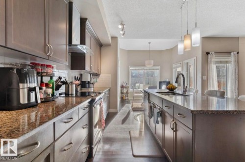 Kitchen featuring dark stone countertops, stainless steel appliances, dark wood finish cabinets, a textured ceiling, and a kitchen island with sink - 21703 86 Avenue, Edmonton, AB - Indoor Photo Showing Kitchen With Upgraded Kitchen