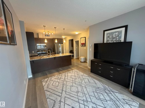Kitchen with a peninsula, light stone counters, tasteful backsplash, stainless steel appliances, and hanging light fixtures - 417 1004 Rosenthal Boulevard, Edmonton, AB - Indoor Photo Showing Living Room