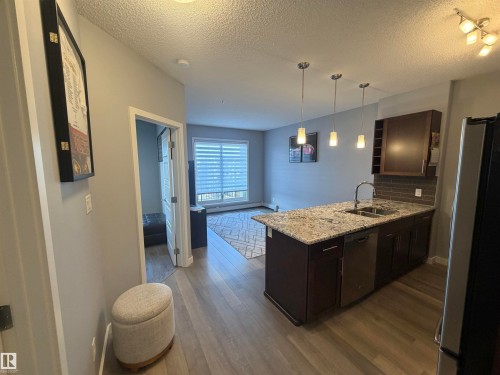 Kitchen with dark wood finish cabinetry, a peninsula, stainless steel appliances, light stone countertops, and dark wood-type flooring - 417 1004 Rosenthal Boulevard, Edmonton, AB - Indoor Photo Showing Kitchen With Double Sink