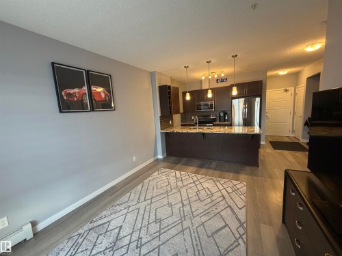 Kitchen featuring dark wood finish cabinetry, light stone counters, a peninsula, a textured ceiling, and stainless steel appliances - 417 1004 Rosenthal Boulevard, Edmonton, AB - Indoor Photo Showing Other Room