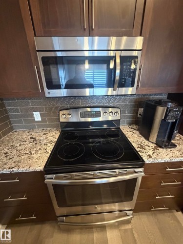Kitchen view of stainless steel appliances, light stone counters, dark wood finish cabinetry, and backsplash - 417 1004 Rosenthal Boulevard, Edmonton, AB - Indoor Photo Showing Kitchen With Upgraded Kitchen