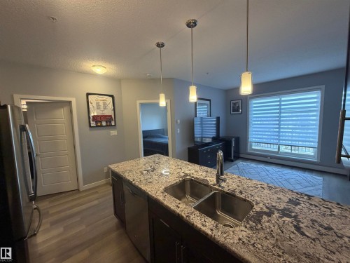 Kitchen with stainless steel appliances, dark wood-type flooring, decorative light fixtures, light stone counters, and a textured ceiling - 417 1004 Rosenthal Boulevard, Edmonton, AB - Indoor Photo Showing Kitchen With Double Sink