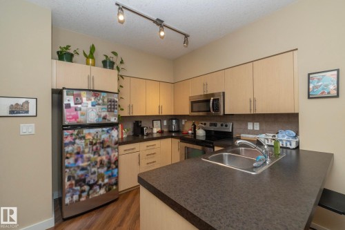 Kitchen featuring stainless steel appliances, dark countertops, a peninsula, light wood finish cabinetry, and rail lighting - 1004 10136 104 Street, Edmonton, AB - Indoor Photo Showing Kitchen With Double Sink
