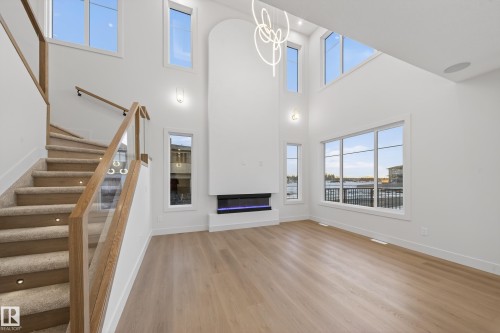 Unfurnished living room featuring light wood-style floors, a glass covered fireplace, hanging lights, and a high ceiling - 8924 Mayday Way, Edmonton, AB 