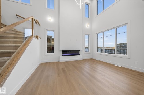 Unfurnished living room featuring light wood-style flooring, a glass covered fireplace, and a high ceiling - 8924 Mayday Way, Edmonton, AB 