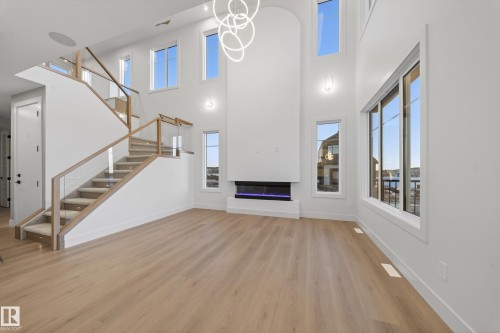 Unfurnished living room featuring light wood-type flooring, hanging lights, a glass covered fireplace, healthy amount of natural light, and a high ceiling - 8924 Mayday Way, Edmonton, AB 