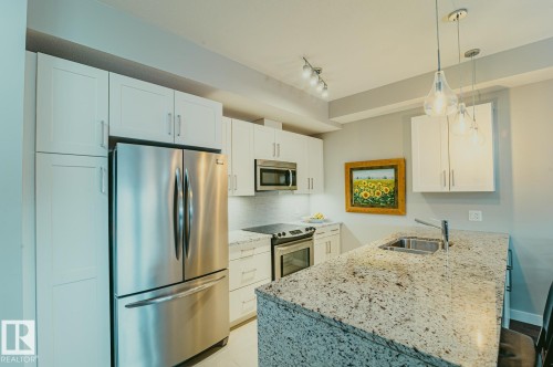 Kitchen featuring stainless steel appliances, a peninsula, solid surface countertops, white cabinets, and pendant lighting - 301 625 Leger Way Nw, Edmonton, AB - Indoor Photo Showing Kitchen With Double Sink With Upgraded Kitchen