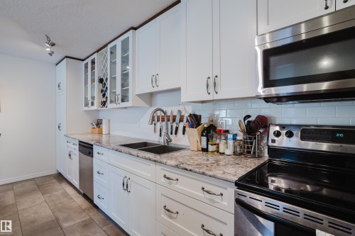 Kitchen with stainless steel appliances, glass insert cabinets, white cabinetry, light stone counters, and a textured ceiling - 115 10545 Saskatchewan Drive, Edmonton, AB - Indoor Photo Showing Kitchen With Double Sink With Upgraded Kitchen