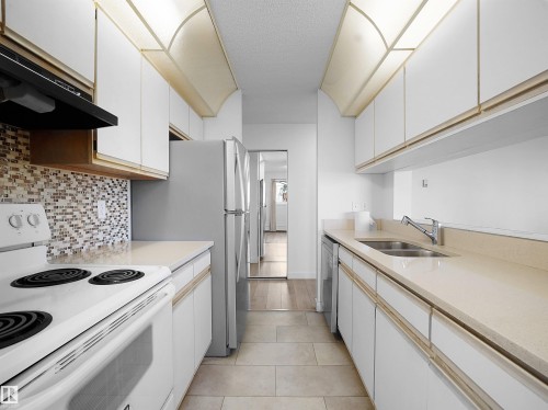 Kitchen with white electric stove, white cabinetry, range hood, backsplash, and a textured ceiling - 108 5065 31 Avenue, Edmonton, AB - Indoor Photo Showing Kitchen With Double Sink