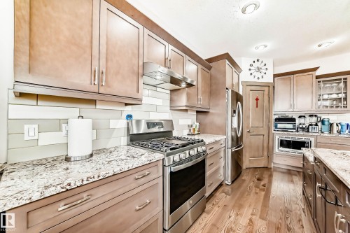 Kitchen featuring stainless steel appliances, glass insert cabinets, light stone counters, light wood-style floors, and tasteful backsplash - 7719 Getty Wynd, Edmonton, AB - Indoor Photo Showing Kitchen With Upgraded Kitchen