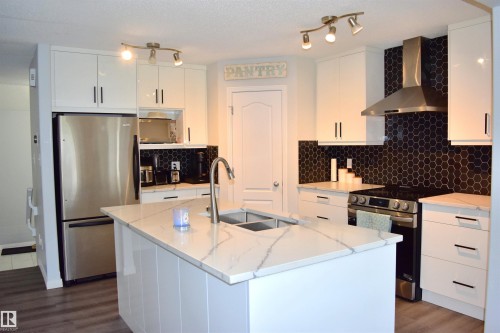 Kitchen with stainless steel appliances, white cabinetry, light stone counters, a kitchen island with sink, and a textured ceiling - 315 Wild Rose Way, Edmonton, AB - Indoor Photo Showing Kitchen With Stainless Steel Kitchen With Double Sink With Upgraded Kitchen