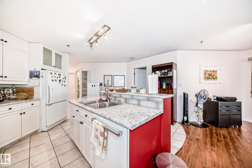 Kitchen with white cabinetry, white appliances, glass insert cabinets, a kitchen island with sink, and light tile patterned floors - 108 12028 103 Avenue, Edmonton, AB - Indoor Photo Showing Kitchen With Double Sink