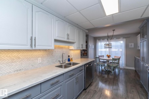 Two tone kitchen featuring decorative light fixtures, stainless steel dishwasher, dark wood-style floors, two tone cabinetry, and light stone countertops - 4371 46 Street, Stony Plain, AB - Indoor Photo Showing Kitchen With Double Sink