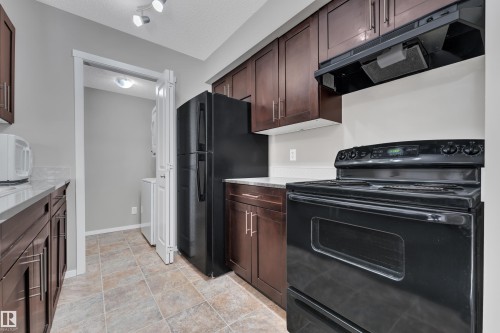 Kitchen featuring black appliances, range hood, a textured ceiling, dark wood finish cabinetry, and rail lighting - 216 5804 Mullen Place, Edmonton, AB - Indoor Photo Showing Kitchen